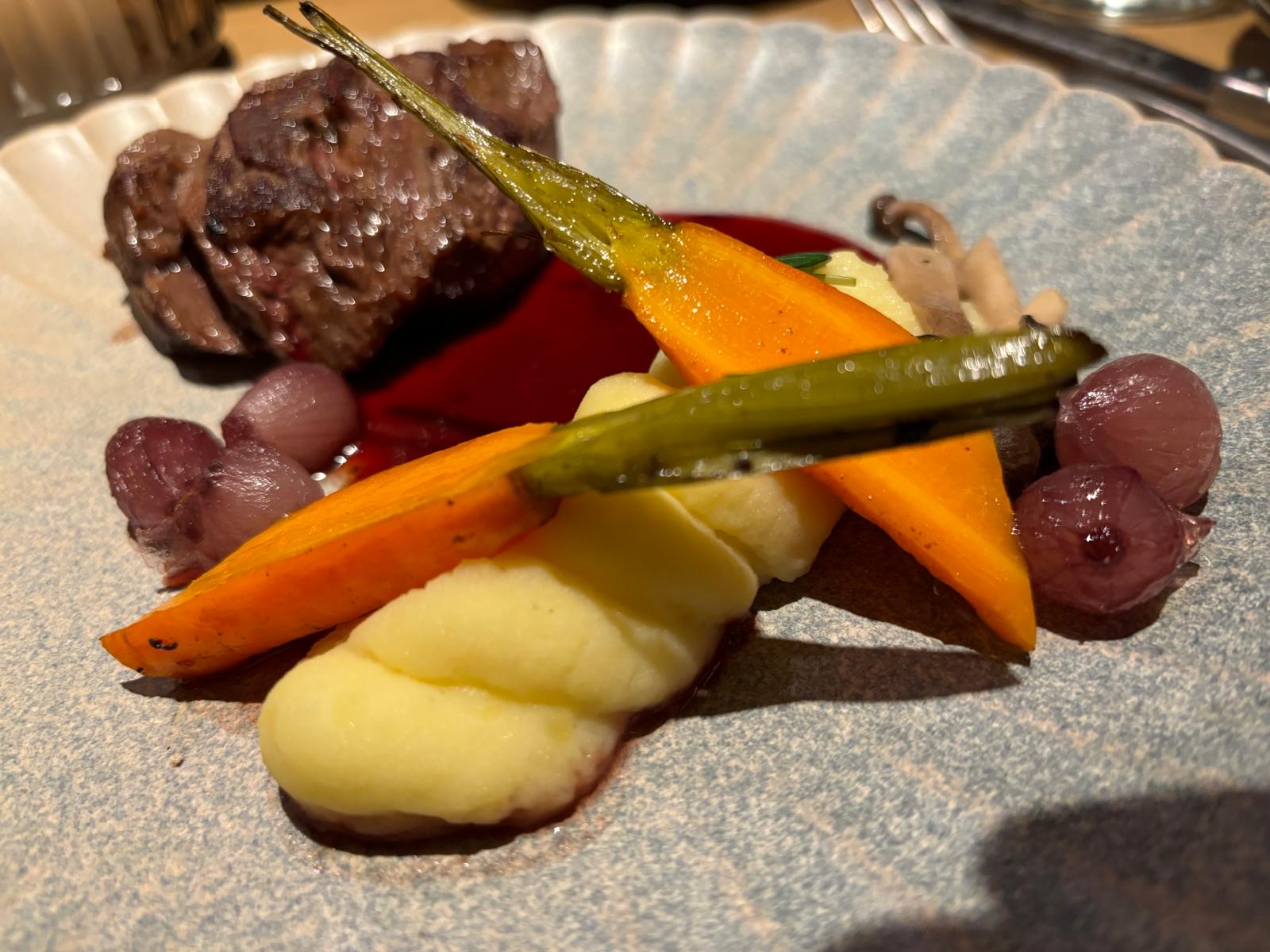 Gebakken vlees met wortel, sjalotjes, aardappelpuree en een rode saus op een licht bord