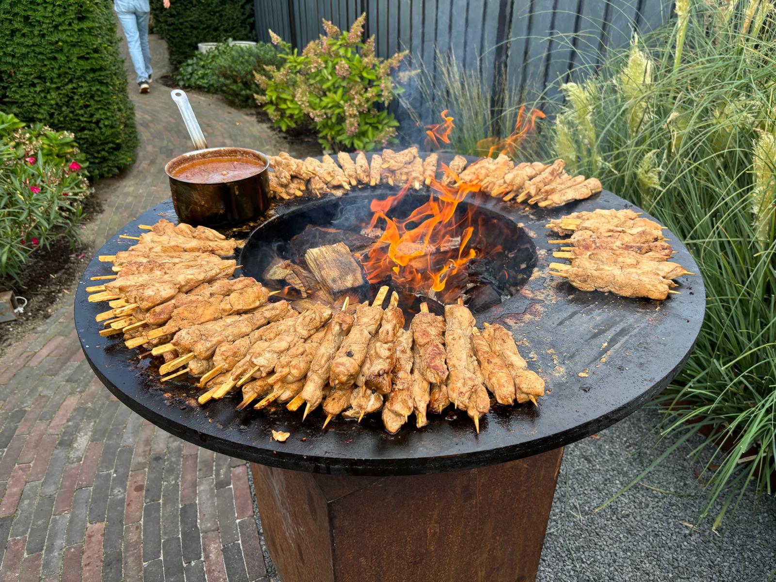 Brandende vuurring in de tuin met rondom tientallen saté-spiesjes en een pan saus